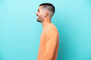 Kafkasyalı genç adam mavi arka planda izole edilmiş. Yan pozisyonda gülüyor.