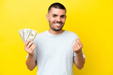 Kafkasyalı genç adam çok para alıyor. Sarı arka planda izole edilmiş. Para hareketi yapıyor.