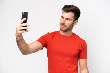 Beyaz arka planda izole edilmiş genç beyaz bir adam cep telefonuyla selfie çekiyor.