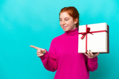 Young caucasian reddish woman holding a gift isolated on blue background pointing to the side to present a product