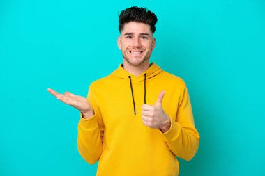 Kafkasyalı genç adam mavi arka planda izole edilmiş. Avucunda hayali bir şey tutuyor. Bir ilan vermek için.