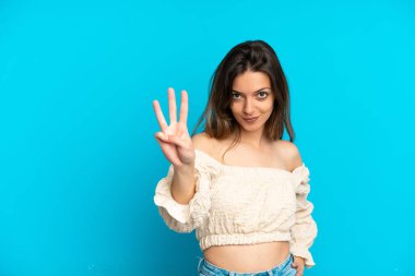 Mavi arka planda izole edilmiş genç beyaz kadın mutlu ve parmaklarıyla üçe kadar sayıyor.