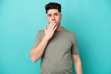 Kafkasyalı genç adam mavi arka planda izole edilmiş esniyor ve geniş açık ağzını eliyle kapatıyor.