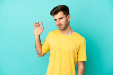 Beyaz tenli yakışıklı adam mavi arka planda izole edilmiş mutlu bir ifadeyle selamlıyor.