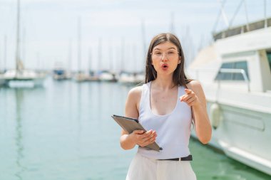 Genç, güzel Ukraynalı bir kadın açık havada tablet tutuyor. Şaşırmış ve ön tarafı işaret ediyor.