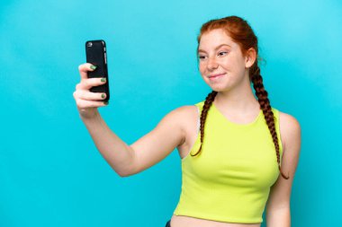 Mavi arka planda izole edilmiş genç kızılımsı bir kadın selfie çekiyor.
