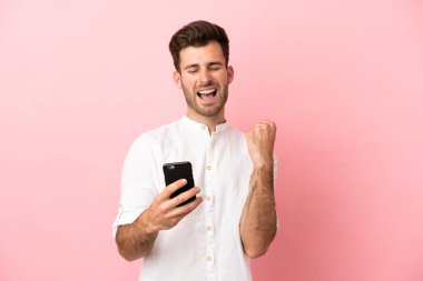 Pembe arka planda izole edilmiş, telefonu zafer pozisyonunda genç beyaz bir adam.