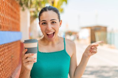 Genç ve güzel bir kadın açık havada kahve tutuyor. Şaşırmış ve parmağıyla işaret ediyor.