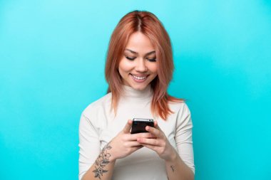 Mavi arka planda izole edilmiş genç bir Rus kadın cep telefonuyla mesaj gönderiyor.