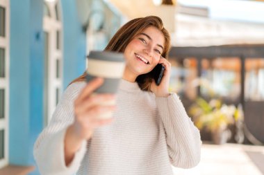 Genç beyaz bir kadın cep telefonu kullanıyor ve elinde mutlu bir ifadeyle kahve tutuyor.