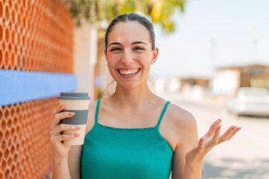 Genç ve güzel bir kadın açık havada kahve tutuyor. Yüzünde şok ifadesi var.