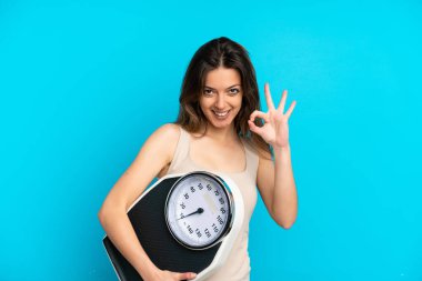 Mavi arka planda izole edilmiş beyaz bir kadın elinde bir tartı makinesi tutuyor ve iyi işareti yapıyor.