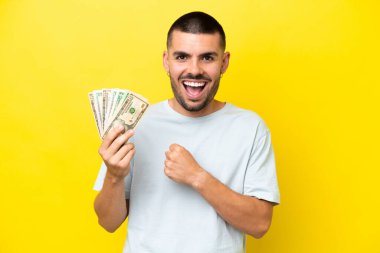 Young caucasian man taking a lot of money isolated on yellow background celebrating a victory
