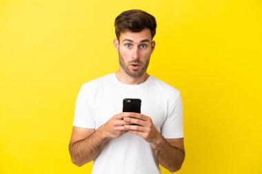 Genç beyaz yakışıklı adam sarı arka planda izole edilmiş kameraya bakarken şaşırmış bir ifadeyle cep telefonunu kullanıyor.