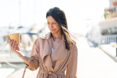 Açık havada elinde kahveyle üzgün bir ifadeyle bekleyen genç bir kadın.