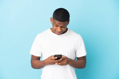 Genç latin adam mavi arkaplanda izole edilmiş cep telefonuyla bir mesaj ya da e-posta gönderiyor