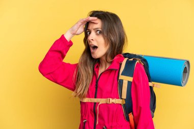 Sarı arka planda izole edilmiş büyük sırt çantalı genç dağcı yan tarafa bakarken sürpriz jest yapıyor.