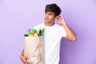Young man holding a grocery shopping bag isolated on purple background having doubts