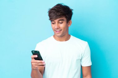 Mavi arka planda izole edilmiş genç beyaz adam cep telefonuyla bir mesaj ya da e-posta gönderiyor