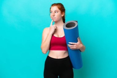 Genç spor kırmızısı kadın yoga derslerine giderken elinde mavi arka planda izole edilmiş bir minder tutarken yukarı bakarken şüphe duyuyor.