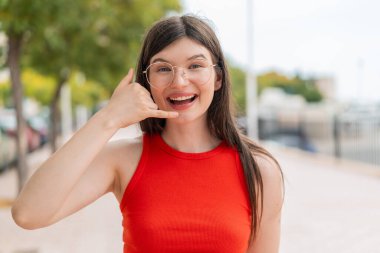 Dışarıda gözlüklü genç bir kadın telefon jesti yapıyor. Bana geri dönüş işareti ver.