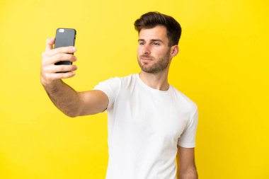 Sarı arka planda izole edilmiş beyaz yakışıklı bir adam cep telefonuyla selfie çekiyor.