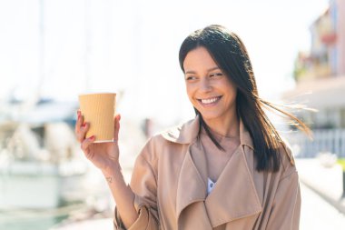Açık havada elinde kahveyle gezen genç bir kadın. Mutlu bir ifadeyle.