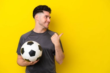Yakışıklı genç futbolcu sarı arka planda izole edilmiş bir şekilde bir ürün sunmak için kenarı işaret ediyor.