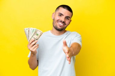 Kafkasyalı genç adam sarı arka planda izole edilmiş bir sürü para alıyor ve iyi bir anlaşma için el sıkışıyor.