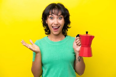 Arjantinli genç bir kadın elinde kahve makinesini tutuyor sarı arka planda izole edilmiş ve şok olmuş bir yüz ifadesi var.