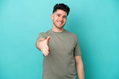 Mavi arka planda izole edilmiş genç beyaz adam iyi bir anlaşma için el sıkışıyor.