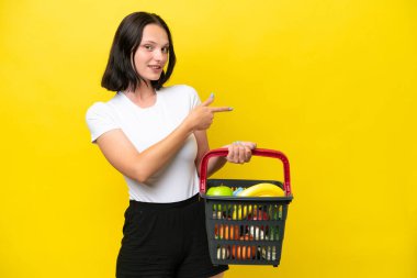 Elinde yiyecek dolu bir alışveriş sepeti olan genç bir kadın sarı arka planda izole edilmiş bir şekilde parmak kaldırıyor.