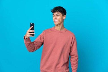 İzole edilmiş mavi arka planı olan genç bir adam selfie çekiyor.