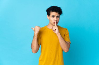 Genç Venezuelalı bir adam mavi arka planda izole edilmiş bir şekilde kenarı işaret ediyor ve sessizlik hareketi yapıyor.