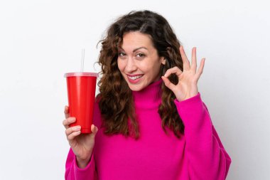 Beyaz arka planda izole edilmiş gazoz içen genç beyaz kadın parmaklarıyla tamam işareti gösteriyor.