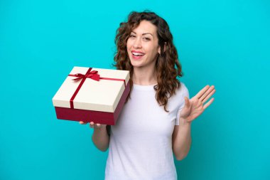Beyaz bir kadın elinde mavi arka planda izole edilmiş bir hediye tutuyor. Mutlu bir ifadeyle selamlıyor.