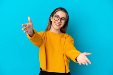 Mavi arka planda izole edilmiş genç beyaz kadın sunar ve davet eder.