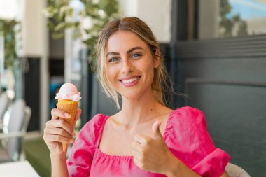 Açık havada kornet dondurmalı genç sarışın kadın. Başparmakları havada. Çünkü iyi bir şey oldu.