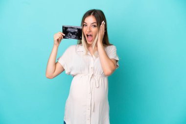 Genç Romen kadın mavi arka planda izole edilmiş hamile ve elinde ultrason tutarken şaşırmış.