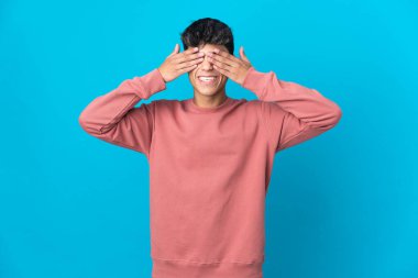 İzole edilmiş mavi arka planda genç bir adam. Gözleri elleriyle kaplıyor ve gülümsüyor.