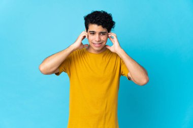 Genç Venezuelalı adam mavi arka planda izole edilmiş hayal kırıklığına uğramış ve kulaklarını kapatmış.