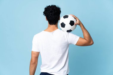 Genç Venezuelalı adam mavi arka planda futbol topuyla izole edilmiş.