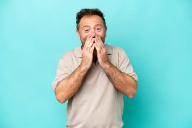 Mavi arka planda izole edilmiş orta yaşlı beyaz bir adam mutlu ve gülümseyen elleriyle ağzını kapatıyor.