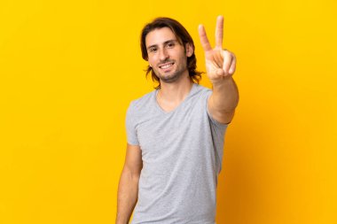 Sarı arka planda saklanmış yakışıklı genç adam gülümsüyor ve zafer işareti gösteriyor.