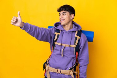Young mountaineer man with a big backpack over isolated yellow background giving a thumbs up gesture
