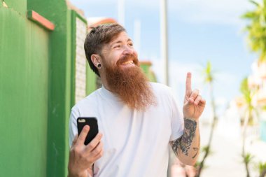 Sakallı kızıl saçlı adam dışarıda cep telefonu kullanıyor. Çözümü bulmak için parmağını kaldırıyor.