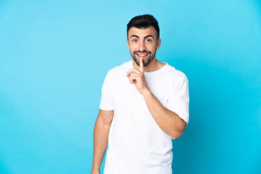 İzole edilmiş mavi arka planda beyaz bir adam sessizlik işareti gösteriyor. Parmağını ağzına sokuyor.