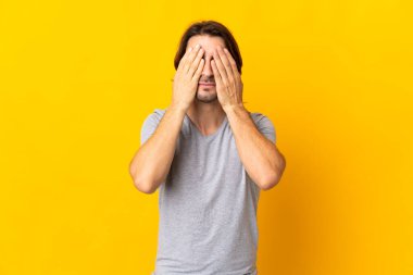 Sarı arka planda izole edilmiş genç yakışıklı adam gözlerini elleriyle kapatıyor.