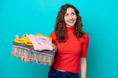 Kafkasyalı genç bir kadın mavi arka planda çamaşır sepetini tutarken yukarı bakarken bir fikir düşünüyor.