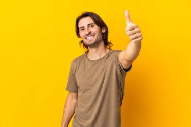 Sarı arka planda izole edilmiş genç ve yakışıklı bir adam. Başparmakları havada. Çünkü iyi bir şey oldu.